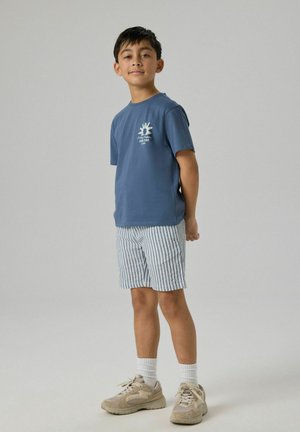 Chłopiec stoi z rękami za plecami, ubrany w niebieską koszulkę z nadrukiem, pasiastymi szortami, białe skarpetki i beżowe trampki na jednolitym tle.