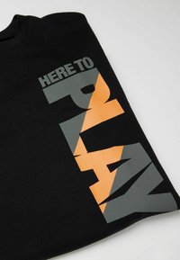 Schwarzes Baumwoll-T-Shirt mit fettem "HERE TO PLAY" Text in Grau und Orange, gestaltet mit einer modernen, kantigen Schriftart.