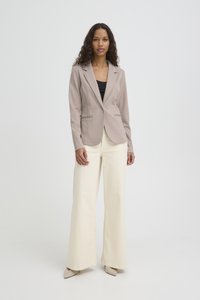 Mujer con cabello largo y rizado que lleva un blazer topo, blusa negra, pantalones crema anchos y zapatos beige puntiagudos, de pie contra un fondo liso.