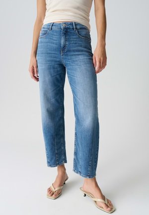 Persona che indossa jeans azzurri a gamba dritta abbinati a sandali beige con tacco e una maglia bianca senza maniche, in piedi davanti a uno sfondo neutro.