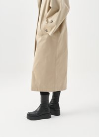 Trench beige con una abertura lateral y acentos de botones, combinado con botas de cuero negro hasta la rodilla con una suela de plataforma gruesa.