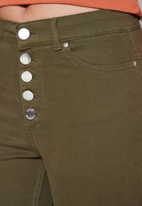 Lähikuva oliivinvihreistä housuista, joissa on viisi hopeista nappia haarassa ja etutasku, yhdistettynä lyhytvartiseen oranssiin toppiin.