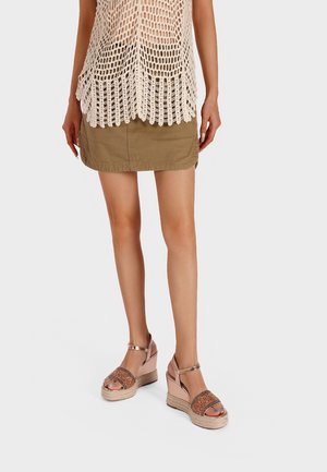 Donna con top beige all’uncinetto, minigonna oliva e sandali con zeppa in oro rosa con cinturini alla caviglia, in piedi su sfondo bianco.