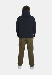 Giacca puffer navy con cappuccio, cuciture visibili e un'etichetta arancione, abbinata a pantaloni cargo oliva. Sneakers bianche completano il look.