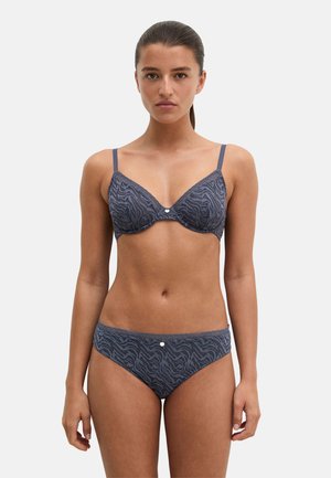 Femme debout face à la caméra, portant un ensemble soutien-gorge et culotte gris à imprimé animalier assorti avec bordure en dentelle sur fond clair uni.