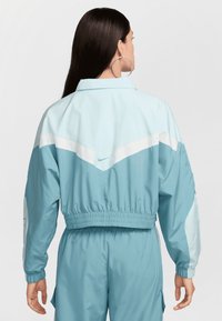 Jachetă bleu deschis cu talie strânsă și manșete, având accente albe într-un model în zigzag. Material fin, ușor, cu tiv elastic.