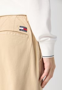 Beżowe spodnie Tommy Jeans z elastycznym pasem i tylną kieszenią, noszone z białą bluzką z długim rękawem, na której widać mankiet i dłoń.