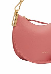 Różowa skórzana torebka na ramię w kształcie półksiężyca, z pozłacanymi elementami i tłoczonym logo. Teksturowana powierzchnia i detal wąskiego mocowania paska.