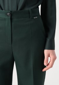 Dark green tailored trousers featuring a smooth texture, a flat front, and a silver "BOSS" logo on the waistband. Button closure visible.