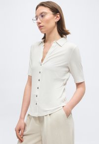 Femeie cu păr scurt și castaniu, purtând ochelari cu rame transparente, o cămașă albă cu mâneci scurte și nasturi, și pantaloni bej deschis, având o mână în buzunar.