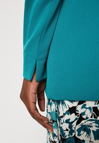 Tyrkysový top s bočními rozparky v kombinaci s květinovou sukní vzoru v černé, bílé a modré barvě. Látka působí hladce a má texturu.