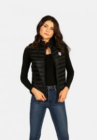 Gilet rembourré noir avec fermeture éclair à l'avant, col montant et motif matelassé. Présente un patch logo sur le côté gauche, associé à un haut noir.