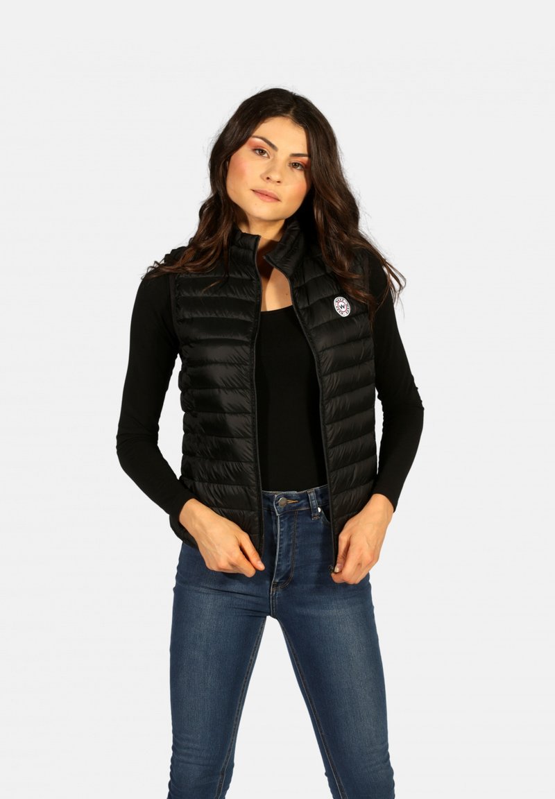 Gilet rembourré noir avec fermeture éclair à l'avant, col montant et motif matelassé. Présente un patch logo sur le côté gauche, associé à un haut noir.