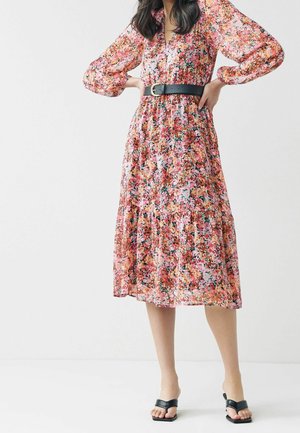 Kukkakuvioinen mekko puff-hihalla, vaaleanpunaisen ja oranssin sävyissä, vyötärölle kiristetty mustalla vyöllä. Kerroksellinen hame, pehmeä tekstuuri ja hyvä virtaus.