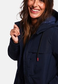 Veste bleu marine avec capuche, dotée d'une fermeture éclair frontale, de boutons-pression et d'un cordon rouge. Texture douce et coupe ajustée.