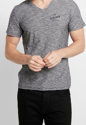 Graues gestreiftes V-Ausschnitt-T-Shirt mit kurzen Ärmeln und einem kleinen Logo nahe der Brust. Kombiniert mit schwarzen Hosen, die Hände vor dem Körper verschränkt.