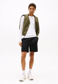 Schwarze sportliche Shorts mit elastischem Bund, kombiniert mit einem weißen Langarmshirt und einem grünen Kapuzensweatshirt, das über die Schultern gelegt ist.