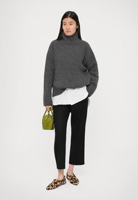 Maglione a collo alto grigio a maglia sopra una camicia bianca, pantaloni neri a vita alta e corti, e pantofole con stampa leopardata, mentre tiene una piccola borsa verde.