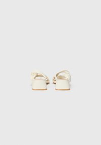 Vue arrière de sandales plates de couleur crème, avec un talon carré, des brides torsadées et une texture lisse avec une semelle marron clair.