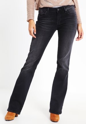 Femme portant un jean bootcut noir, un pull en maille beige et des bottines en daim beige, debout jambes légèrement écartées.