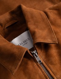 Bruine suède jas met een ritsluiting, puntige kraag en een label met de tekst "LES DEUX." Close-up toont de zachte textuur en het zilveren ritsdetail.