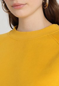 Sudadera amarilla con cuello redondo acanalado y tejido suave, que presenta mangas raglán y sin patrones ni acentos visibles.