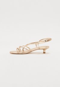 Sandales strappy métalliques dorées avec un petit talon kitten. Elles sont dotées de lanières croisées, d'une bride de cheville réglable et d'une semelle lisse et plate.