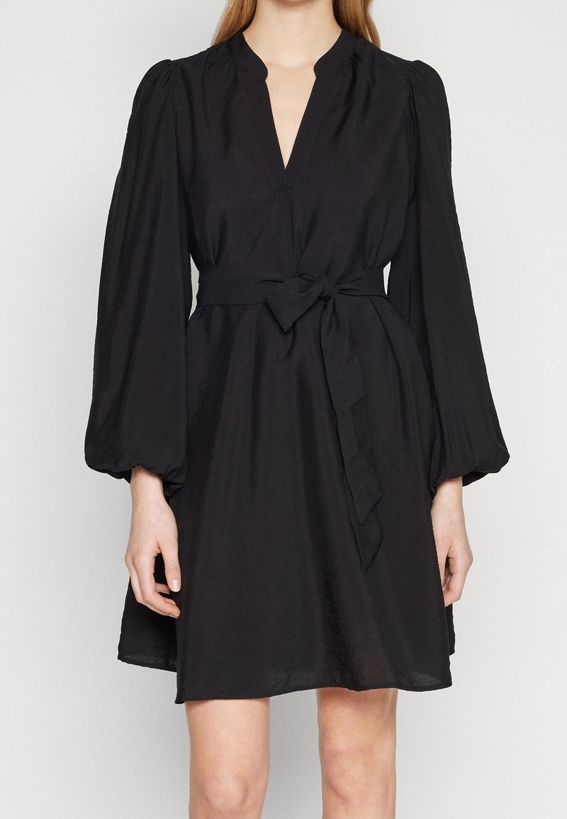 Femme portant une robe noire longueur genou avec de longues manches bouffantes, un décolleté en V et une ceinture en tissu nouée à la taille.
