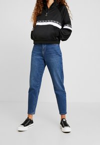 Černá zipová mikina s bílou a černou logovou páskou, zkombinovaná s vysokým pasem modrými džíny a černými teniskami s bílou podrážkou.