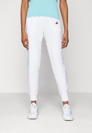 Personne portant un pantalon de survêtement Adidas blanc, des baskets blanches et un haut bleu clair, debout devant un fond uni clair.