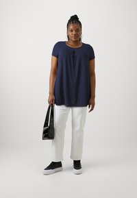 Marineblauw shirt met korte mouwen en een ronde hals, met een losse pasvorm, gecombineerd met witte wijde broek en zwarte sneakers, met een zwarte handtas in de hand.