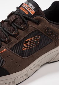 Brązowe i czarne buty sportowe, wykonane z siateczki i zamszu, z pomarańczowymi akcentami i teksturowaną gumową podeszwą. Widoczne logo marki.