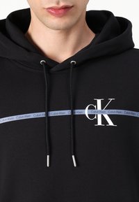 Crna dukserica s kapuljačom napravljena od pamuka. Sadrži zatvaranje s vezicom, srebrne detalje i plavi logo Calvin Klein na traci preko grudi.