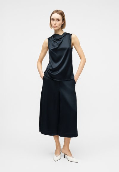 Γυναίκα που φορά ασύμμετρο μαύρο σατέν τοπ χωρίς μανίκια και μαύρο παντελόνι με φαρδιά μπατζάκια μέχρι τον αστράγαλο, με λευκά μυτερά τακούνια, στέκεται μπροστά σε απλό φόντο.