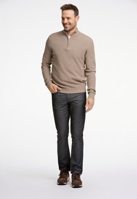 Suéter de manga larga color marrón claro con cremallera de cuarto y punto texturizado; combinado con unos vaqueros oscuros ajustados y zapatillas deportivas marrones. Un atuendo sencillo y funcional.