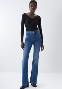 Top lungo a maniche nere a coste con dettagli in pizzo e bottoni, abbinato a jeans a campana blu con cinque tasche e vita alta.