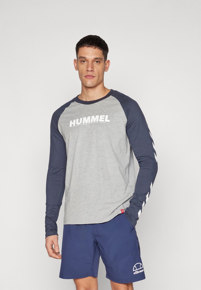 Μακρύ μανίκι μπλούζα με ανοιχτό γκρι σώμα και ναυτικές μανίκια, που παρουσιάζει το "HUMMEL" σε έντονη λευκή γραμματοσειρά στο στήθος και σχήματα βέλους στα μανίκια.