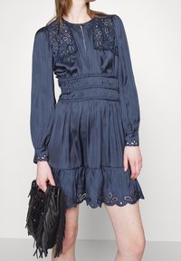 Femme portant une robe bleu marine à manches longues avec des détails en dentelle sur le col, les poignets et l'ourlet, tenant un sac à main noir à franges dans la main gauche.