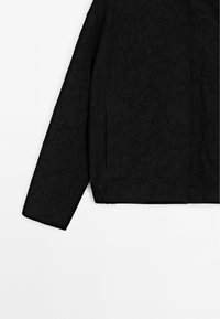Veste noire à manches longues avec un motif floral discret, un ourlet élastique et une poche fendue verticale sur le côté.