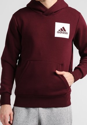 Mężczyzna w bordowej bluzie z kapturem Adidas z przednią kieszenią i białą naszywką z logo, w połączeniu z jasnoszarymi spodniami dresowymi.