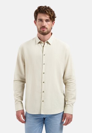 Jonge man met kort krullend bruin haar, gekleed in een beige overhemd met lange mouwen en lichtblauwe spijkerbroek, tegen een witte achtergrond.