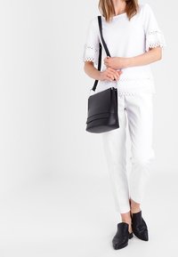 Černá crossbody taška s texturovaným designem a vrstvami na přední straně; nosí se s bílým topem s krajkovým okrajem, bílými kalhotami a černými muly.