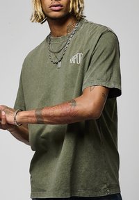 T-shirt en coton vert olive à manches courtes, avec un petit logo graphique sur la poitrine et une coupe décontractée. Manches avec poignets repliés et texture remarquable.