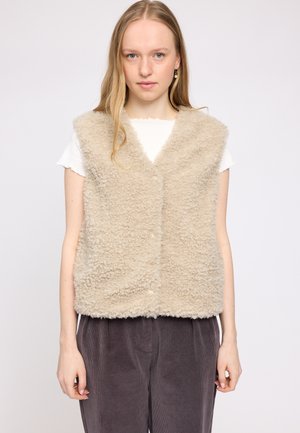 Beige, flauschige Weste mit V-Ausschnitt, Knopfverschluss vorne und ohne Ärmel, über einem weißen Hemd getragen, kombiniert mit dunkelbraunen Cordhosen.