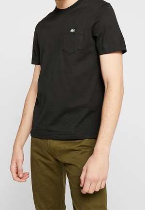 Homme portant un t-shirt noir à manches courtes avec logo sur la poche poitrine et un pantalon vert olive, debout devant un fond clair uni.