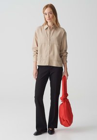 Beige overhemd met knopen en een kraag, gecombineerd met zwarte flared broek. Model houdt een felrode, zachte handtas vast. Eenvoudig, modern ontwerp.