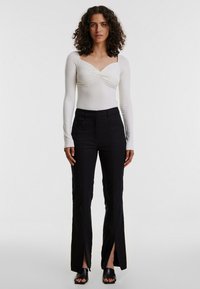 Weißes Langarmoberteil mit geknoteter Vorderseite, kombiniert mit schwarzen hochgeschnittenen Hosen mit Seitenschlitzen und glatter Textur. Schwarze offene High Heels.