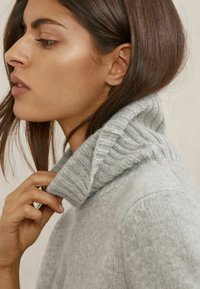 Maglione dolcevita grigio a coste con collo spesso, caratterizzato da una texture morbida. Il modello tira delicatamente il collo con una mano.