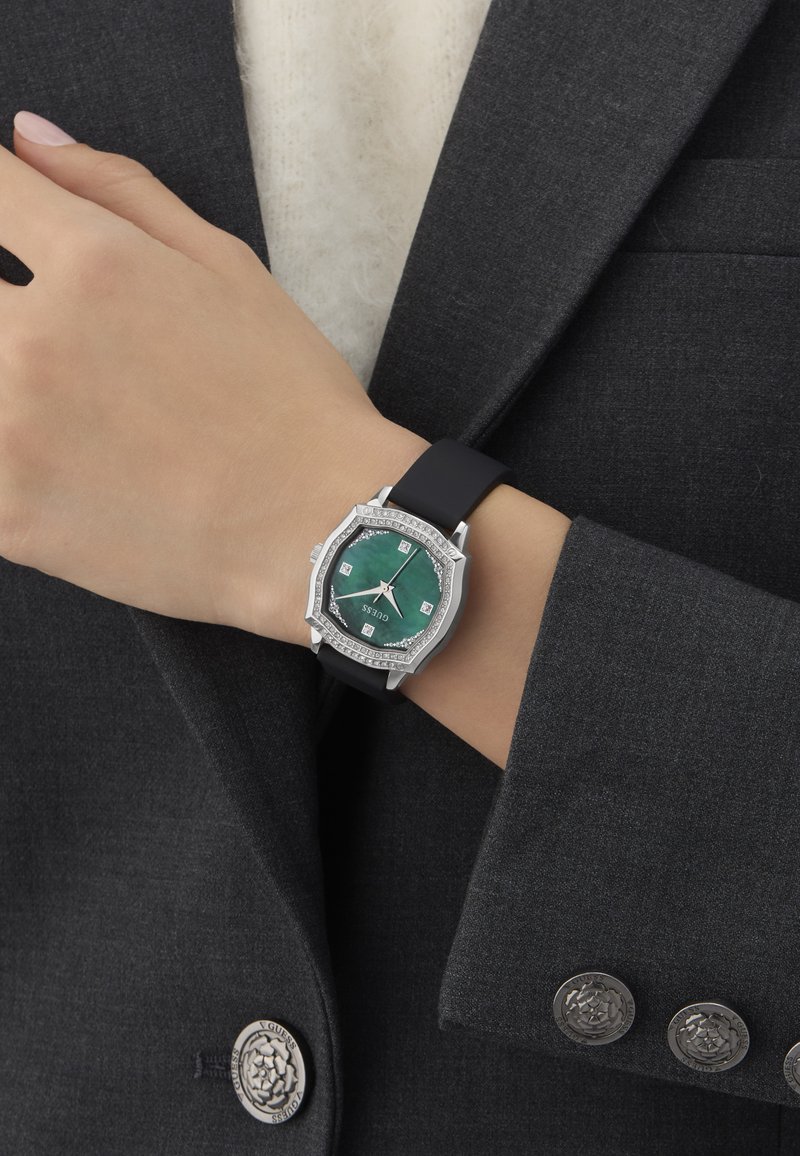 Rand must valge musta rihmaga kell, millel on roheline kaheksanurkne nägu ja kristallaktsendid, kantud tumeda halli bleiseri varruka peal.