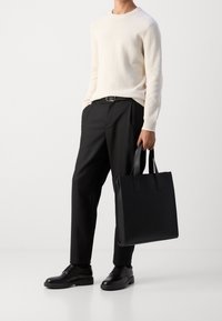 Schwarze Leder-Shopper-Tasche mit zwei oberen Griffen, rechteckige Form, glatte Textur. Getragen von einer Person in einem cremefarbenen, gerippten Pullover und schwarzen Hosen.